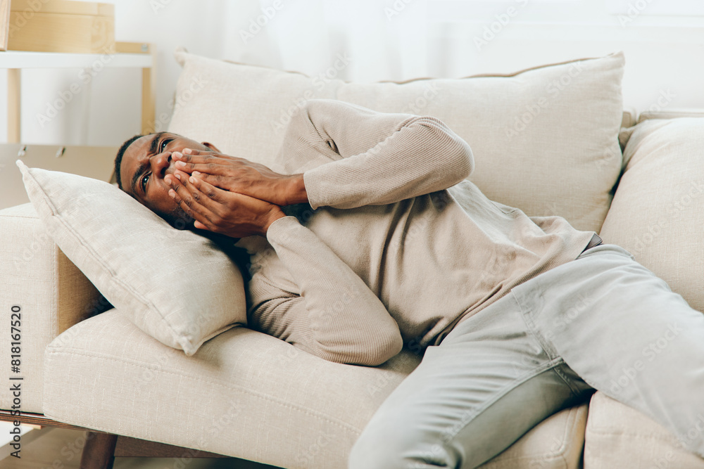 Obraz premium Stressed African American Man with Headache Sitting on Couch at Home The man, exhausted and sicklooking, clutches his head in pain as he rests on the sofa in his living room The combination of