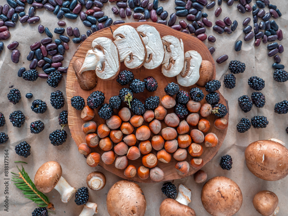 Healthy autumn food selection: white sliced mushrooms, shelled hazelnuts, purple kidney beans, fresh blackberries and tiny fir branch on a slice of wood. Top view, flat lay.