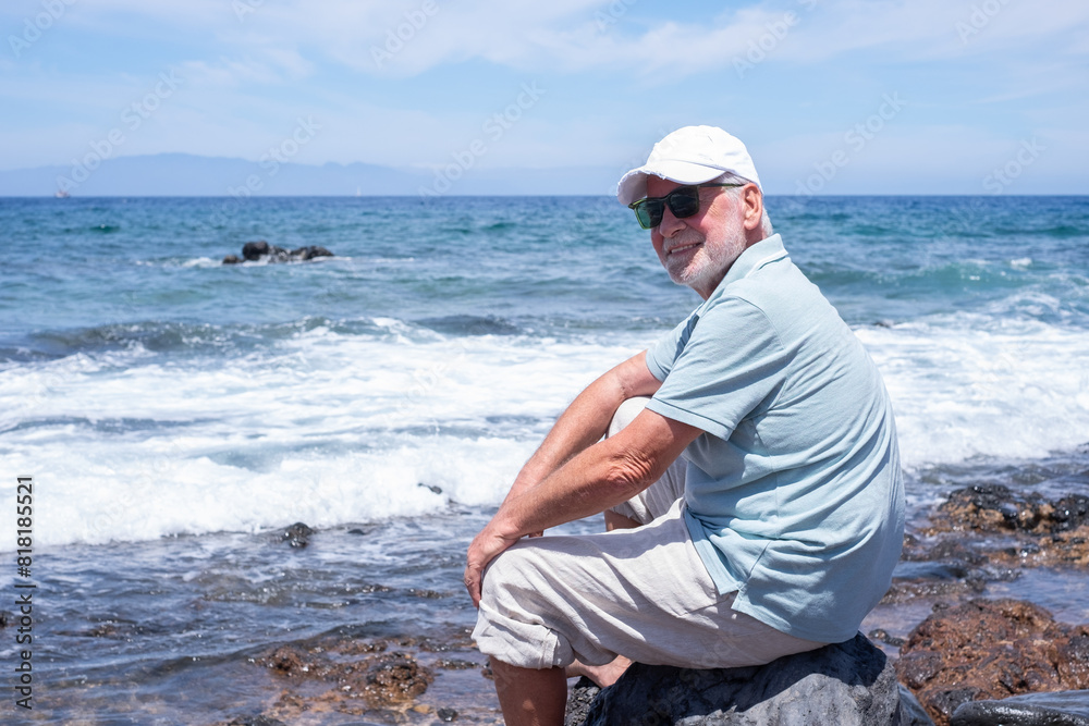Carefree senior relaxed man barefoot wearing sunglasses and cap sitting on a rocky beach admiring the sea waves. Travel vacation leisure activity concept