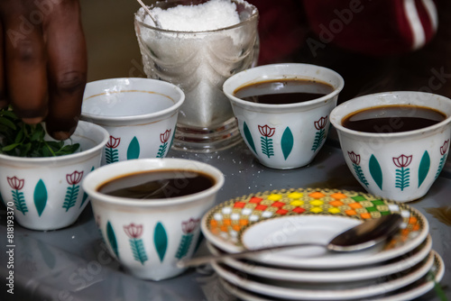 A jebena sits on a grass-covered table with small, handle-less cups arranged around it. Roasted coffee beans, a mortar and pestle, and incense smoke create a traditional Ethiopian coffee ceremony