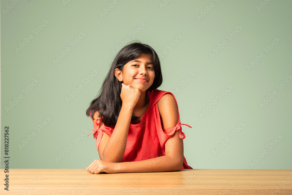 Indian asian small girl sitting at table against plain green background Indian asian small girl sitting at table against plain green background
