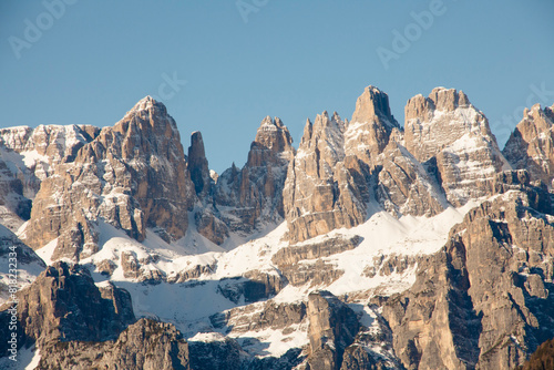 The stunning mountain Paganella in Italy, with rugged, jagged peaks partially covered in snow. Steep rock faces and snow-dusted ridges. Majestic landscape under a clear blue sky.
