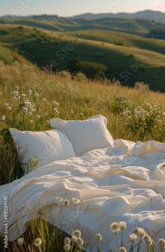 A cozy white bed with pillows and a comforter set up outdoors in a lush meadow, surrounded by wildflowers and rolling green hills. photo