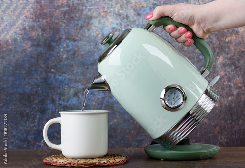 A woman pours hot water from a green electric kettle into a light-colored cup. © svdolgov
