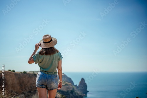 A woman wearing a straw hat stands on a cliff overlooking the ocean