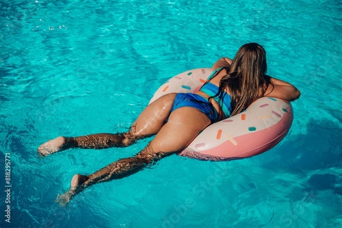 A woman is floating on a pink donut shaped float in a pool. The pool is blue and the woman is wearing a blue bikini. © svetograph