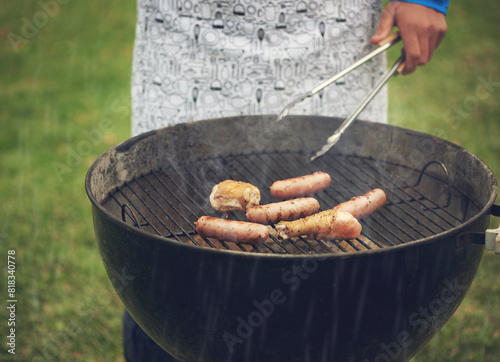 Outdoor, grill and hand for cooking, person and food on fire for barbecue, backyard and nature of grass. Home, lawn and apron for meal prep, meat and hungry for eating, rain, sausage or morning