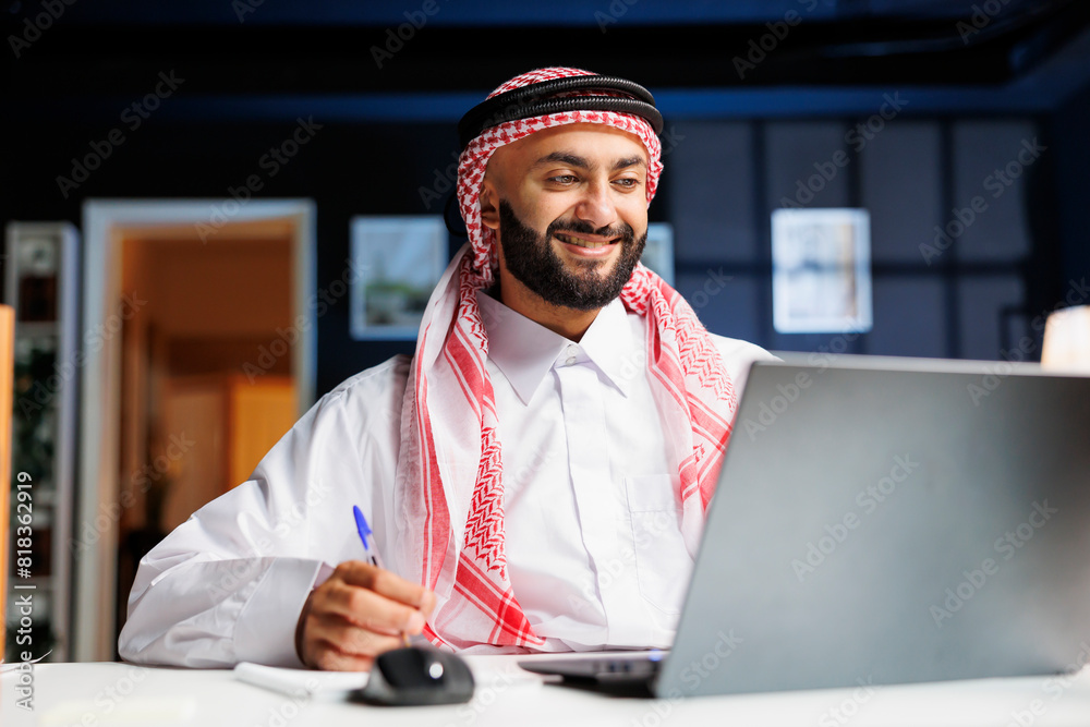 Smiling Arab guy works diligently at office desk, engages in digital tasks and writes on his notepad with laptop nearby. He efficiently conducts online research, embodying dedication and productivity. Smiling Arab guy works diligently at office desk, engages in digital tasks and writes on his notepad with laptop nearby. He efficiently conducts online research, embodying dedication and productivity.