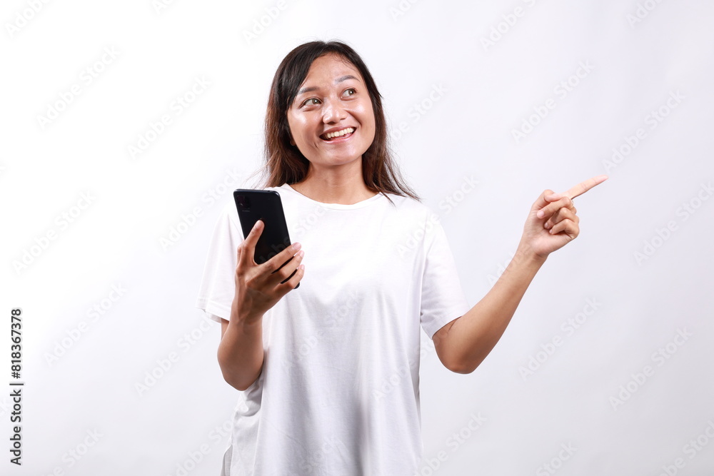 Excited young asian woman shopping on mobile app, holding phone and pointing finger left at empty copy space, showing promo text, standing with smartphone against white background Excited young asian woman shopping on mobile app, holding phone and pointing finger left at empty copy space, showing promo text, standing with smartphone against white background