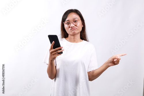 Skeptical asian modern girl, holding smartphone, pointing finger left at empty space and frowing hesitant, has doubts. Isolated on white background photo