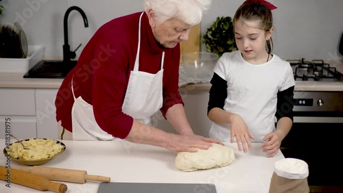 Elderly woman teach cute girl to cook. Kid help to granny knead dough on kitchen table. Spend time at home with children cooking together. Family cookery share skills knowledge and experience