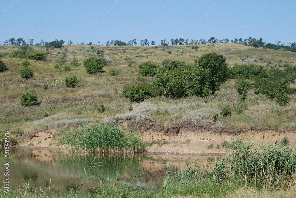 Hillside on the bank of a small river. Summer day in the countryside. On the slope of a high hill grass grows, trees and bushes have green leaves. The grass was partially burned out by the sun. Hillside on the bank of a small river. Summer day in the countryside. On the slope of a high hill grass grows, trees and bushes have green leaves. The grass was partially burned out by the sun.