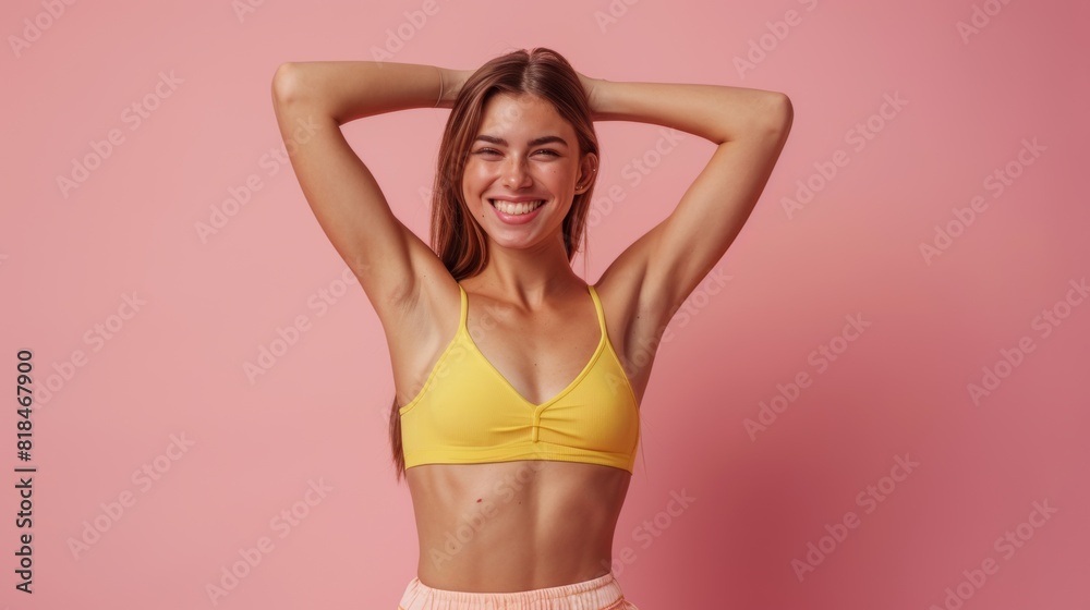 Naklejka premium Smiling woman in yellow sports bra with hands behind head on a pink background, expressing happiness and confidence.