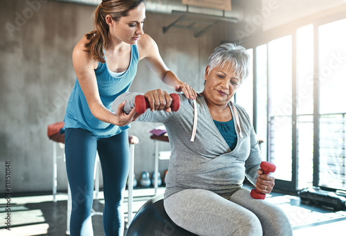 Senior woman, weights and helping with coach in gym for physio, arm exercise and rehabilitation on ball. Old female patient, instructor and workout equipment for arthritis with balance and dumbbells photo