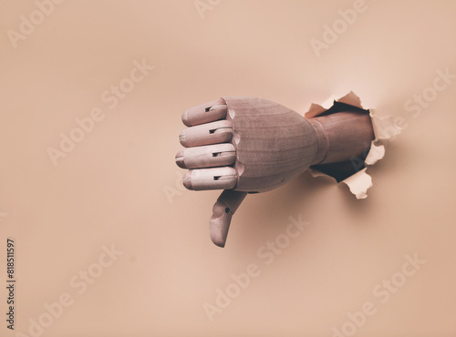 A right wooden hand protrudes from a torn hole in yellow paper and shows a thumbs down (dislike). The concept of disapproval and that things will go badly. Artificial intelligence, robot. Monochrome.