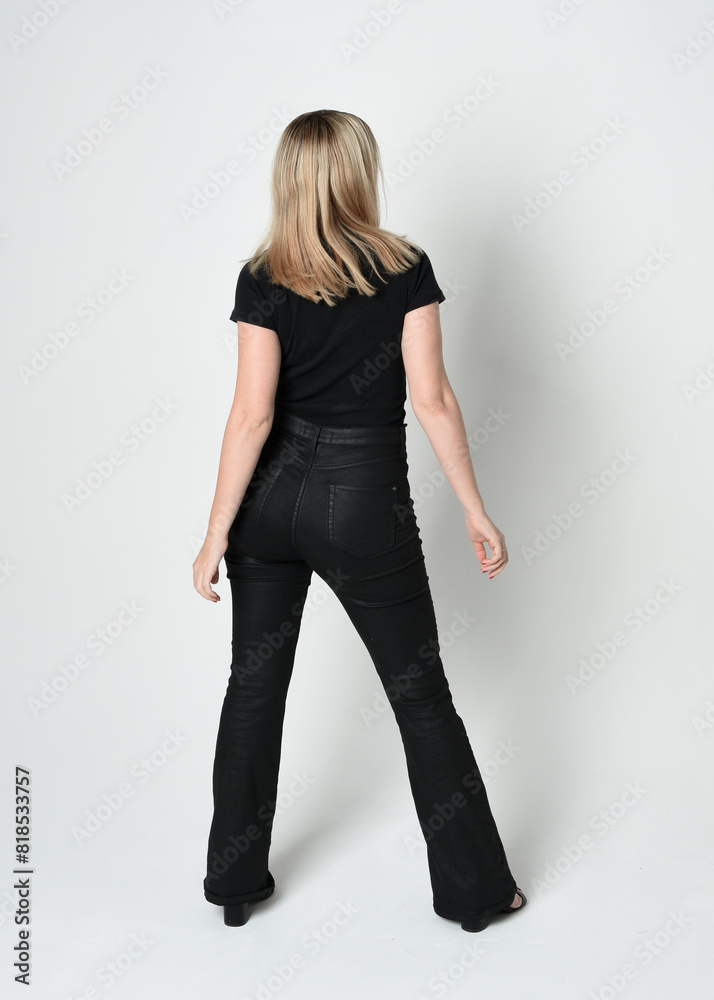 Full length portrait of beautiful blonde woman wearing modern black shirt and leather pants. Confident standing pose walking away from camera, silhouetted on white studio background. Full length portrait of beautiful blonde woman wearing modern black shirt and leather pants. Confident standing pose walking away from camera, silhouetted on white studio background.
