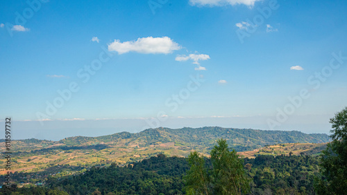 The image offers a panoramic view of a hilly landscape under a vast, clear blue sky. The hills roll gently across the terrain, adorned with patches of greenery and brown earth, suggesting a mix of veg