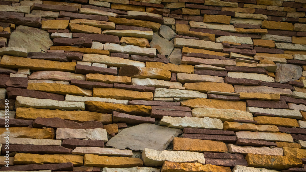  close-up view of a stone wall composed of irregularly shaped stones in various shades of brown, grey, and beige. The stones are arranged in a staggered pattern, giving the wall an intricate and rusti