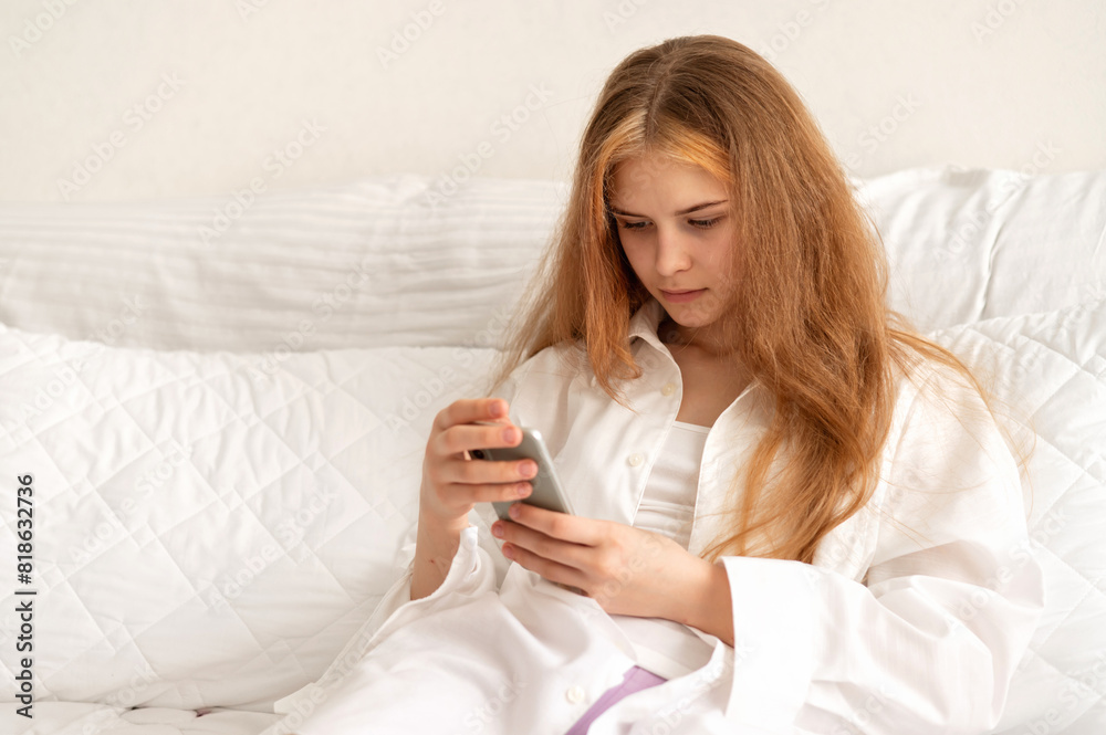Young teenage girl wearing white shirt sitting in neutral white bedroom with smartphone in hands looking on mobile screen Young teenage girl wearing white shirt sitting in neutral white bedroom with smartphone in hands looking on mobile screen