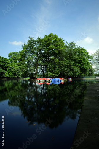 Long exposure of red and cream pedalo boats parked up under lush green trees at a boating lake. One is draped with a blue tarpaulin