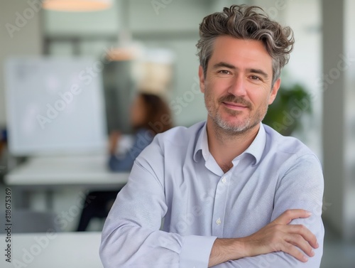 A confident businessman with a friendly smile poses in a modern office setting. The blurred background features a colleague working, emphasizing a professional and collaborative environment.