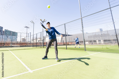A girl in sportswear is training on a paddle tennis court. The girl is hitting the ball against the glass to make a rebound. Concept of women playing paddle. © Angelov