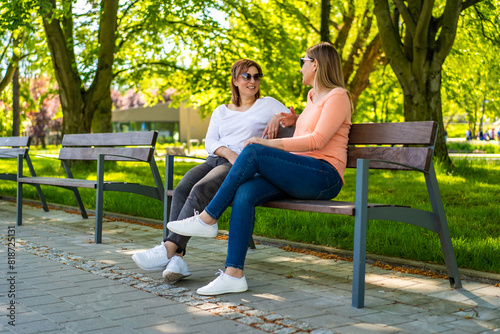 Two mid-adult beautiful women sitting on bench in city park and talking