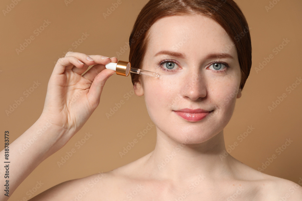 Beautiful woman with freckles applying cosmetic serum onto her face on beige background