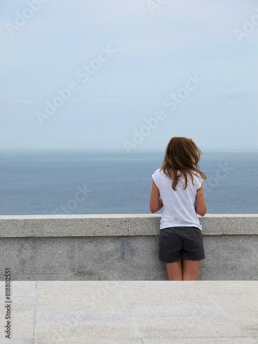 woman with her back looking at the sea, from a stone railing, with her hair blown by the sea breeze. Image in blue tones