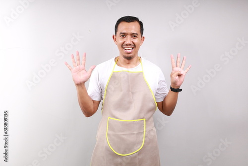 Happy Asian male housekeeper chef cook baker man wear apron showing nine fingers isolated on gray background studio Cooking food concept photo