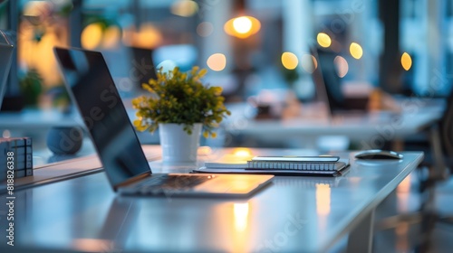A laptop, notebook, pen, and a plant on a desk with blurred office background and warm lights