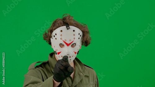 A maniac in a hockey mask and jumpsuit points his finger at the camera. Unrecognizable man in scary Jason Voorhees guise on green screen chromakey. Halloween, horror.