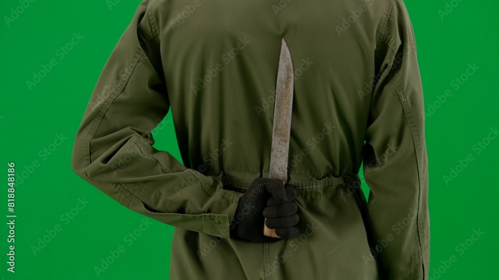 Fototapeta premium Rear view of a maniac hiding a knife behind his back. Man in jumpsuit with weapon on green screen chroma key close up. Halloween, horror.