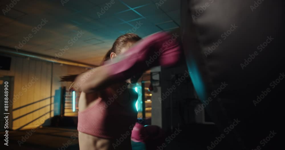 Young woman throwing a powerful punch at a heavy bag in a dimly lit gym with an illuminated boxing ring, showcasing intense training and fitness dedication Young woman throwing a powerful punch at a heavy bag in a dimly lit gym with an illuminated boxing ring, showcasing intense training and fitness dedication