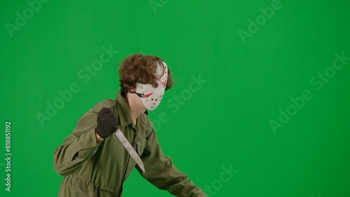 Maniac in hockey mask and jumpsuit running and waving a large sharp knife. An unrecognizable man in the terrifying guise of Jason Voorhees on a green screen chromakey. Halloween, horror. Side view.