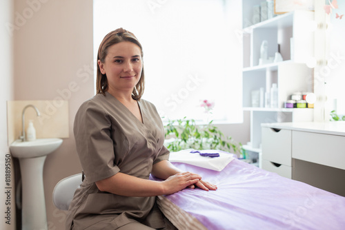 Cheerful young female cosmetologist in beige uniform sitting in her light white office. Skin therapist, beautician and skincare professional concept