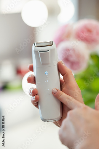 Professional device for face massage in hand of cosmetologist, close up, pink flowers on the background. Tool for body and skin care, spa procedure concept