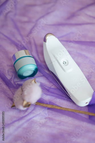 Ultrasonic skin scrubber, small glass container and one cotton stem lying on the table, covered with violet veil, close up, copy space. Concept of professional devices, body and skin care