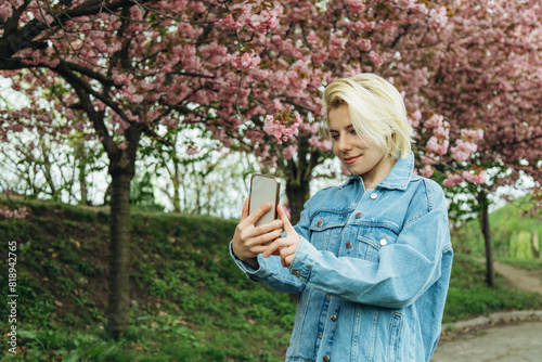 A young blonde woman takes a selfie against the background of cherry blossoms on her phone on a sunny spring day.