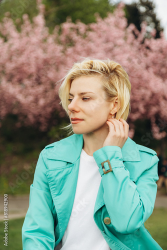 A young dreamy woman in a turquoise trench coat fixes her blond hair while enjoying the cherry blossoms in the park.