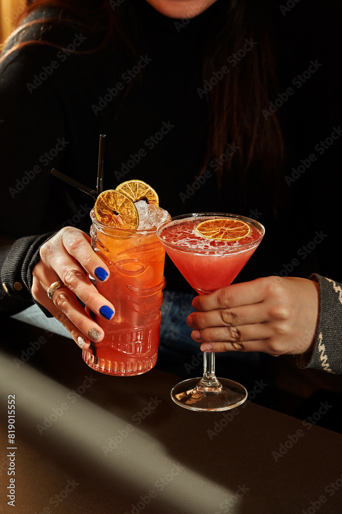 © nazarovsergey - Women clinking glasses with colorful cocktails in dimly lit bar