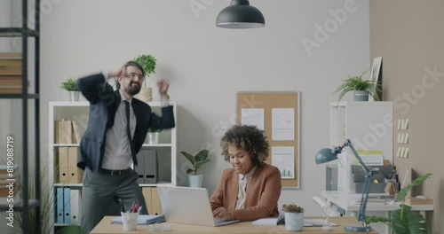 African American businesswoman working with laptop computer while Middle Eastern businessman dancing in office. Workplace and business activity concept. photo