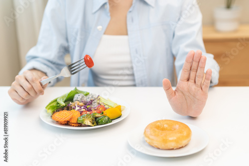 Diet concept, close up young woman, girl using hand push out, stop sweet donut, dessert or junk food on plate, choose green vegetables salad, eat low fat for good health. Female getting weight loss. © KMPZZZ