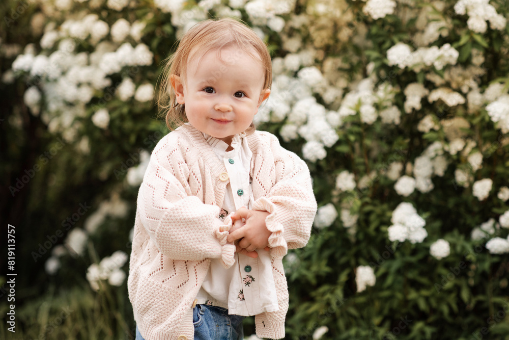 Smiling baby girl 1-2 year old wearing stylish clothes standing over blooming bushes with flowers outdoor. Looking at camera. Childhood.