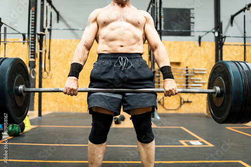 An unrecognizable senior man performs shirtless physical exercise inside a gym. The male is lifting a bar with metal discs to work his legs. Deadlift concept in older men.