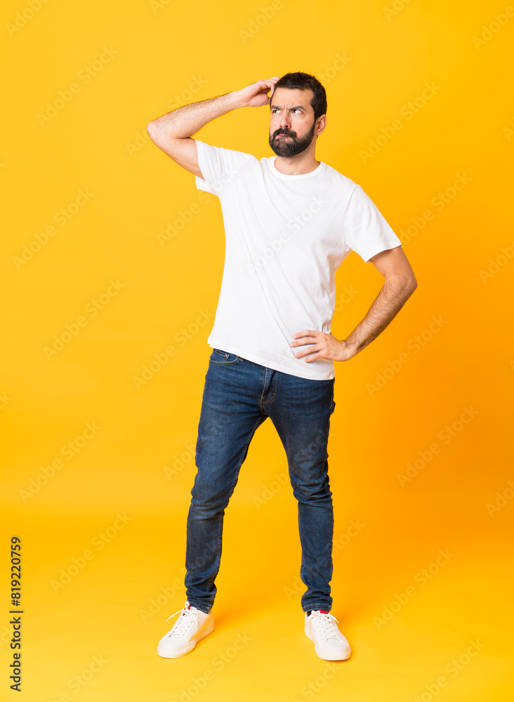 Full-length shot of man with beard over isolated yellow background having doubts while scratching head Full-length shot of man with beard over isolated yellow background having doubts while scratching head