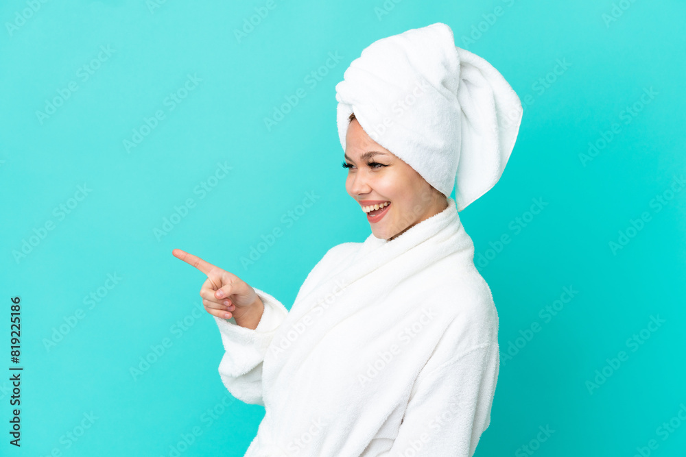 Teenager blonde girl in a bathrobe over isolated blue background pointing finger to the side and presenting a product Teenager blonde girl in a bathrobe over isolated blue background pointing finger to the side and presenting a product