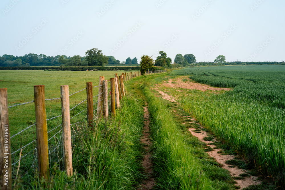 Leading lines along a wooden fence in the British countryside. Public footpath with agriculture fields, crops and farmland. Beautiful English landscape on warm sunny day in spring summer. Leading lines along a wooden fence in the British countryside. Public footpath with agriculture fields, crops and farmland. Beautiful English landscape on warm sunny day in spring summer.