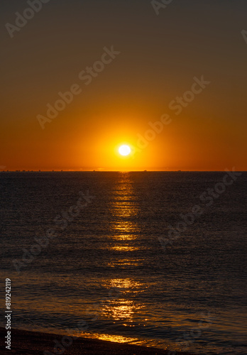 Calm sea illuminated by the orange light of the bright dawn sun illuminating the silhouettes of a group of fishing boats fishing for Palamos shrimp in the Mediterranean Sea.
