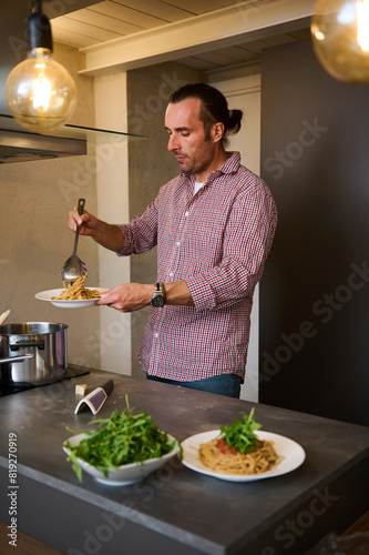 Caucasian handsome chef cooking Italian pasta at home kitchen, pouring tomato sauce over freshly boiled spaghetti. Fresh ingredients on the countertop. Culinary. Epicure. Traditional Italian cuisine.